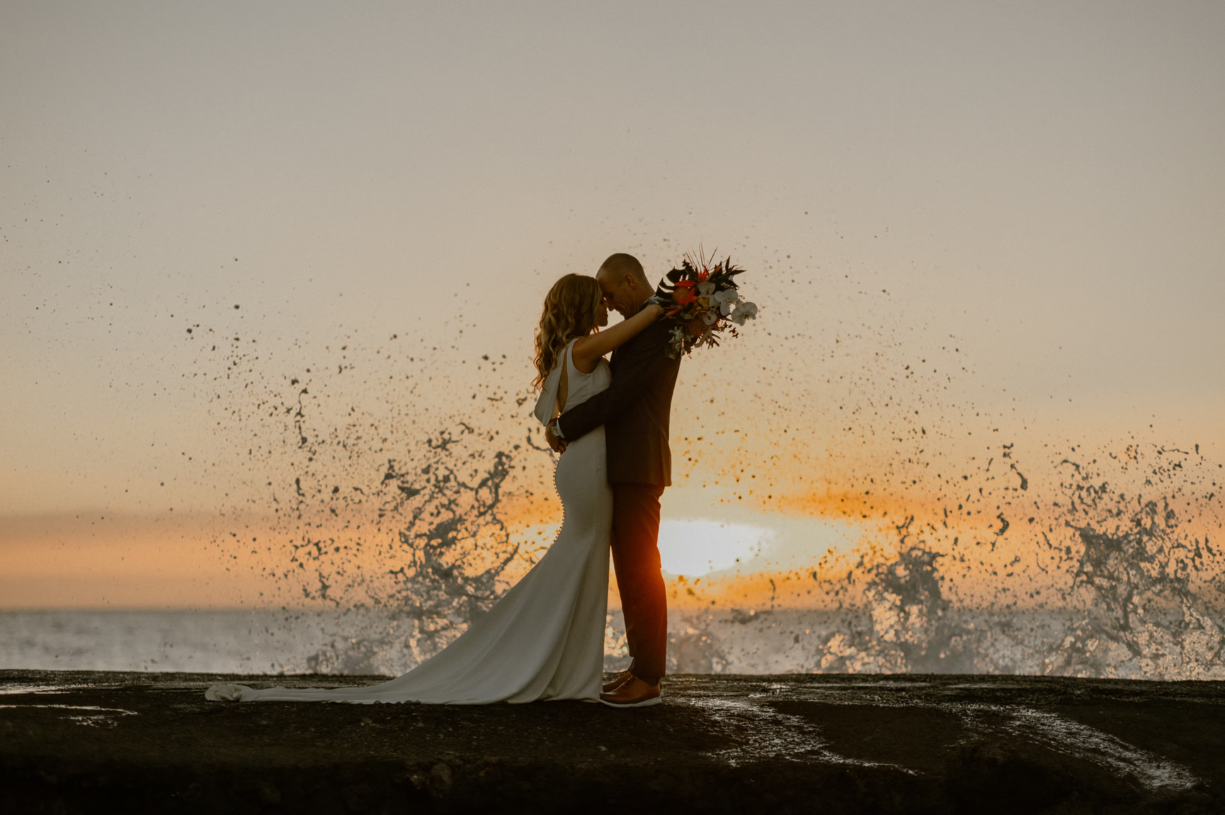 Silhouetted couple in formal wedding attire embrace on lava rock at sunset, a wave breaking high into a spray of water and light behind them, the orange and grey sky of late dusk filling the upper half of the frame.