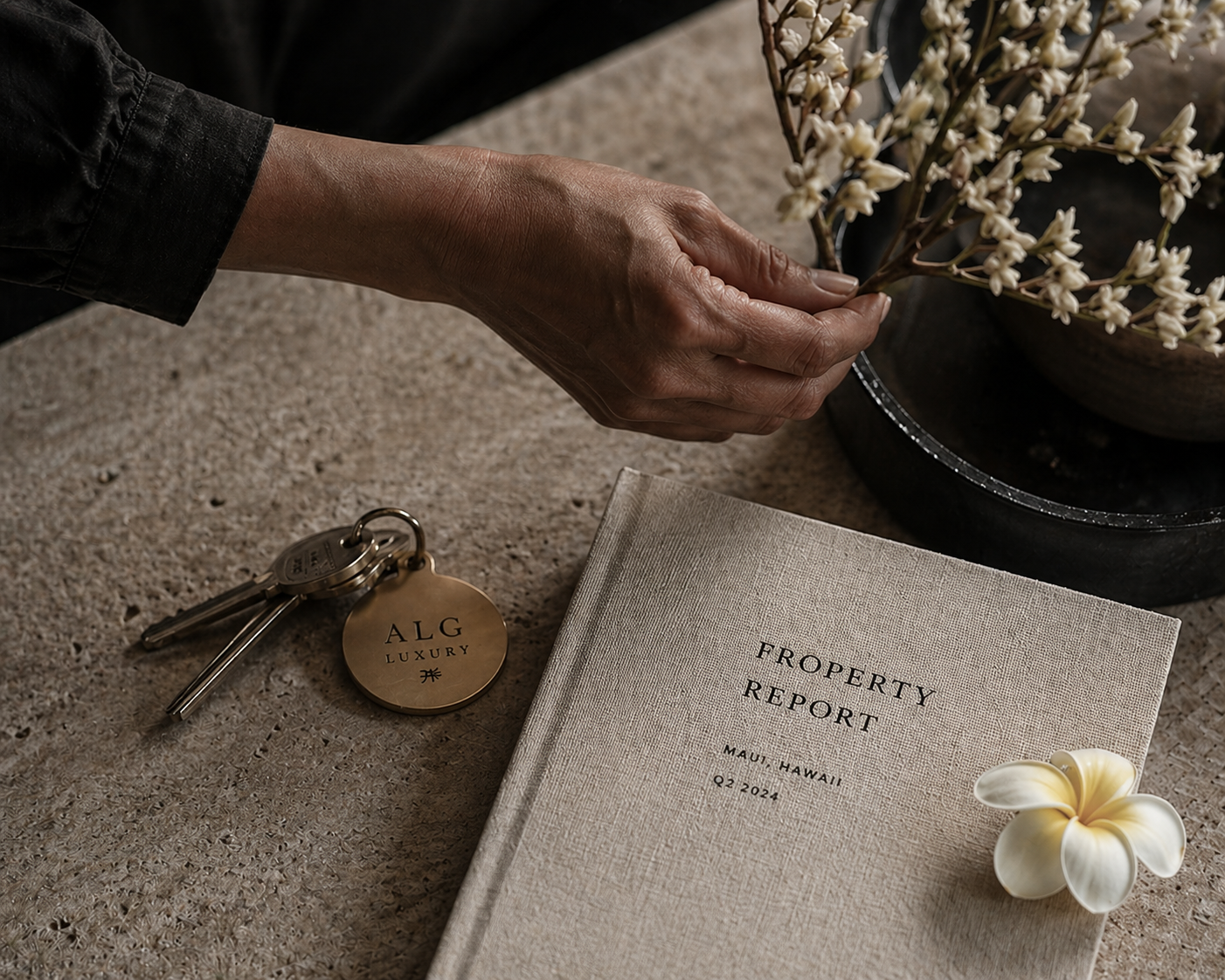 A hand arranging dried botanical stems in a stone vessel beside a linen-bound property report and a brass ALG Luxury keychain, late afternoon light across the stone surface.